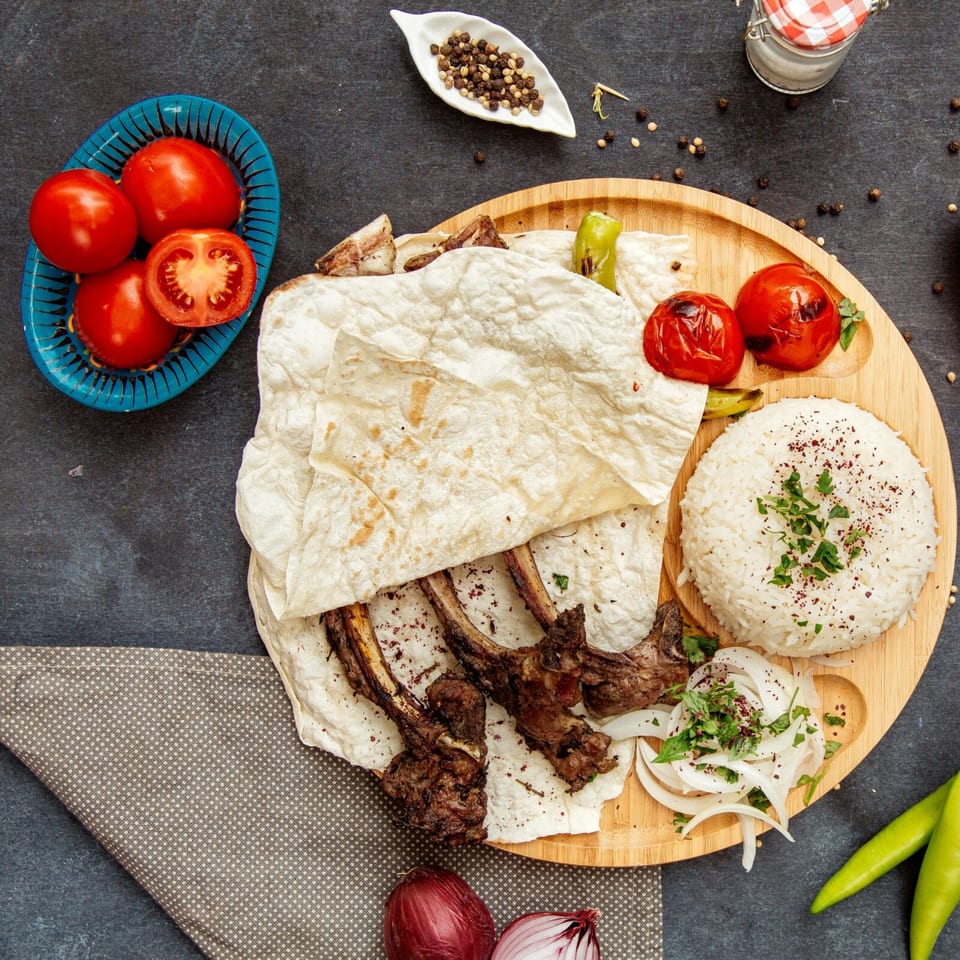 Lebanese lamb chops with rice, flatbread, grilled tomatoes, and onions on a wooden platter.