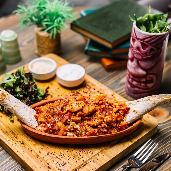 Lebanese chicken shawarma in a clay dish with flatbread, salad, and sauces on a wooden board.