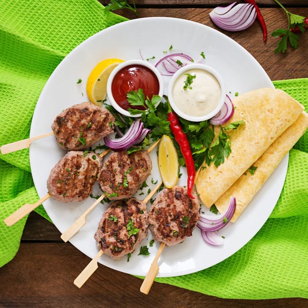 Lebanese kofta kebabs with sauces, lemon, red onion, chili, parsley, and flatbreads on a white plate.